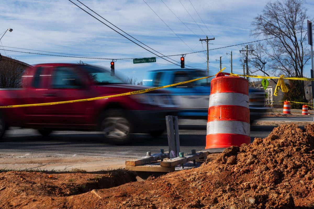Cars zoom by the construction of a pedestrian crosswalk at the intersection of University Parkway and Long Drive.