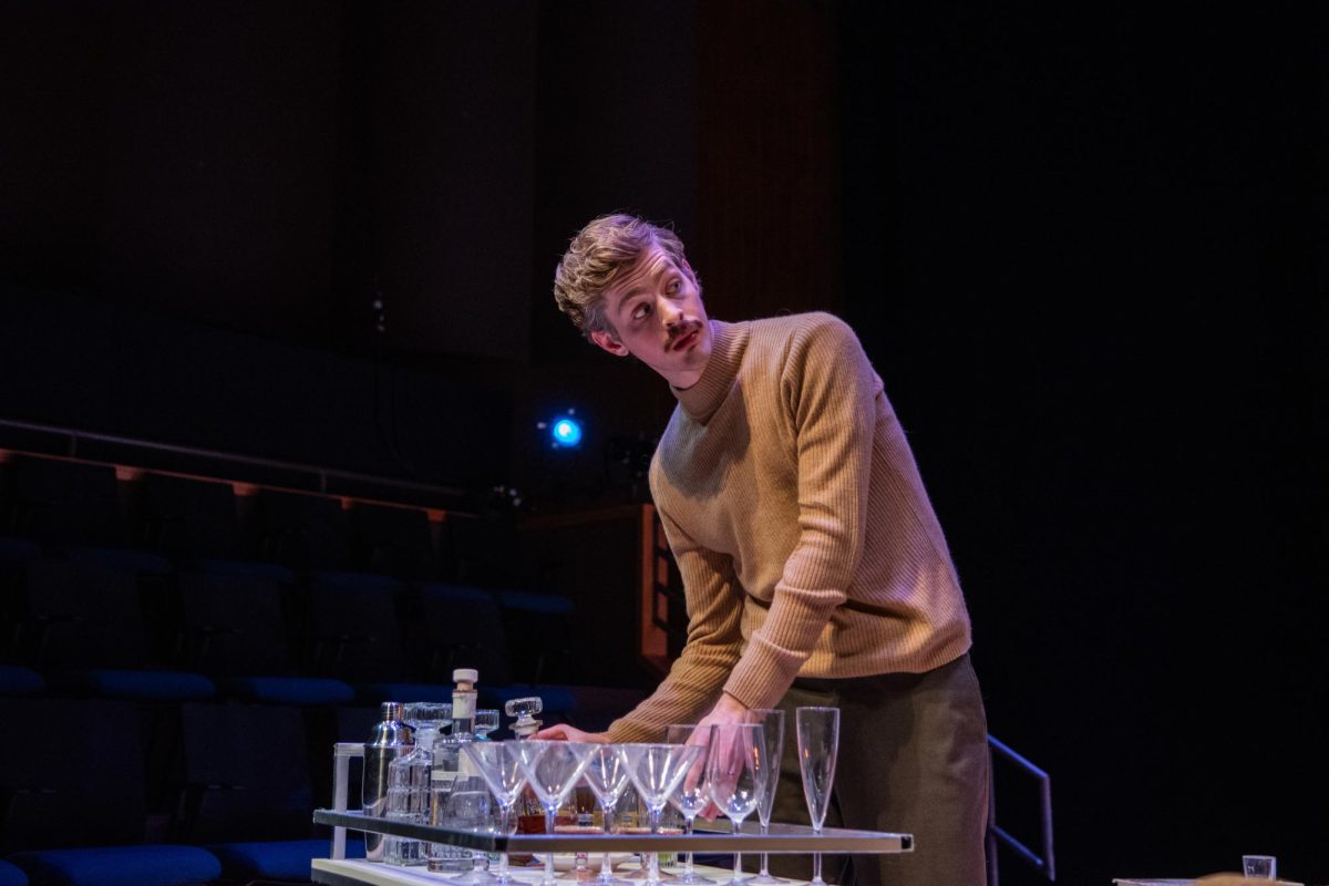 Bennet Haara, playing Deeley, pours himself a drink as he glances back at his fellow cast members.